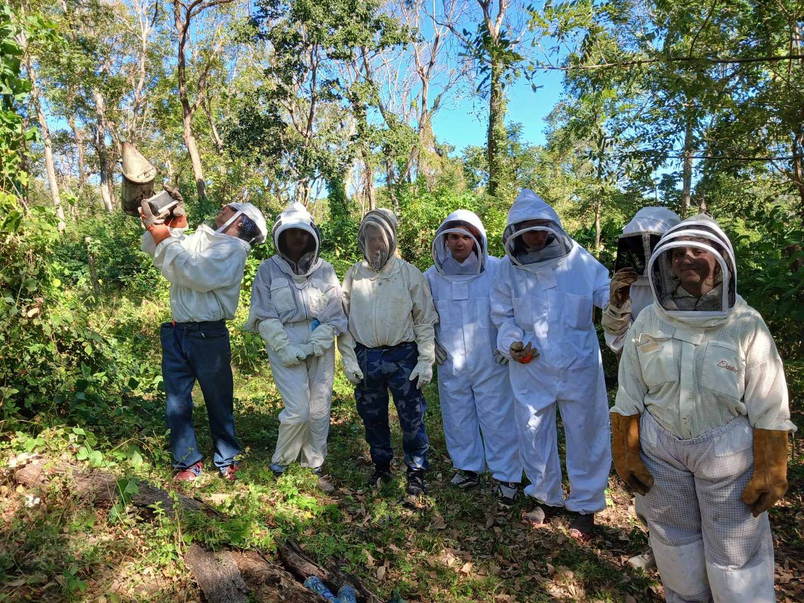 A group in full beekeeper gear gathered in the forest with a local beekeeper using a smoker