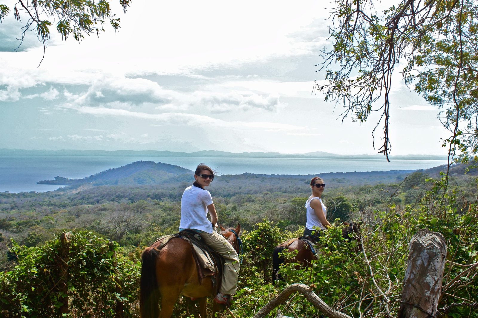 Two riders on horseback overlooking a vast panorama of jungle canopy and Lake Nicaragua from a hilltop mirador